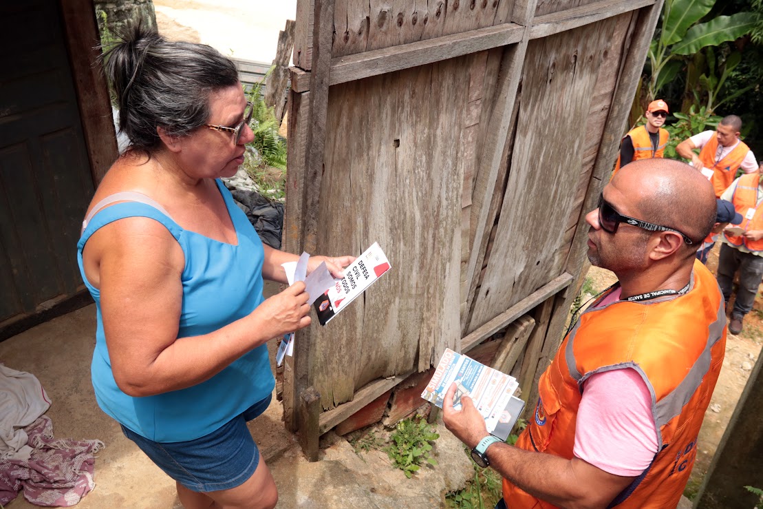 Agente da Defesa Civil entregando folheto de orientação para moradora na Vila Progresso em Santos