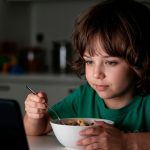 Criança utiliza celular durante a refeição enquanto faz uma refeição à mesa