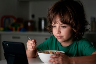 Criança utiliza celular durante a refeição enquanto faz uma refeição à mesa