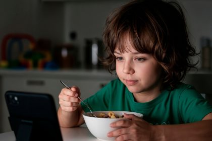 Criança utiliza celular durante a refeição enquanto faz uma refeição à mesa
